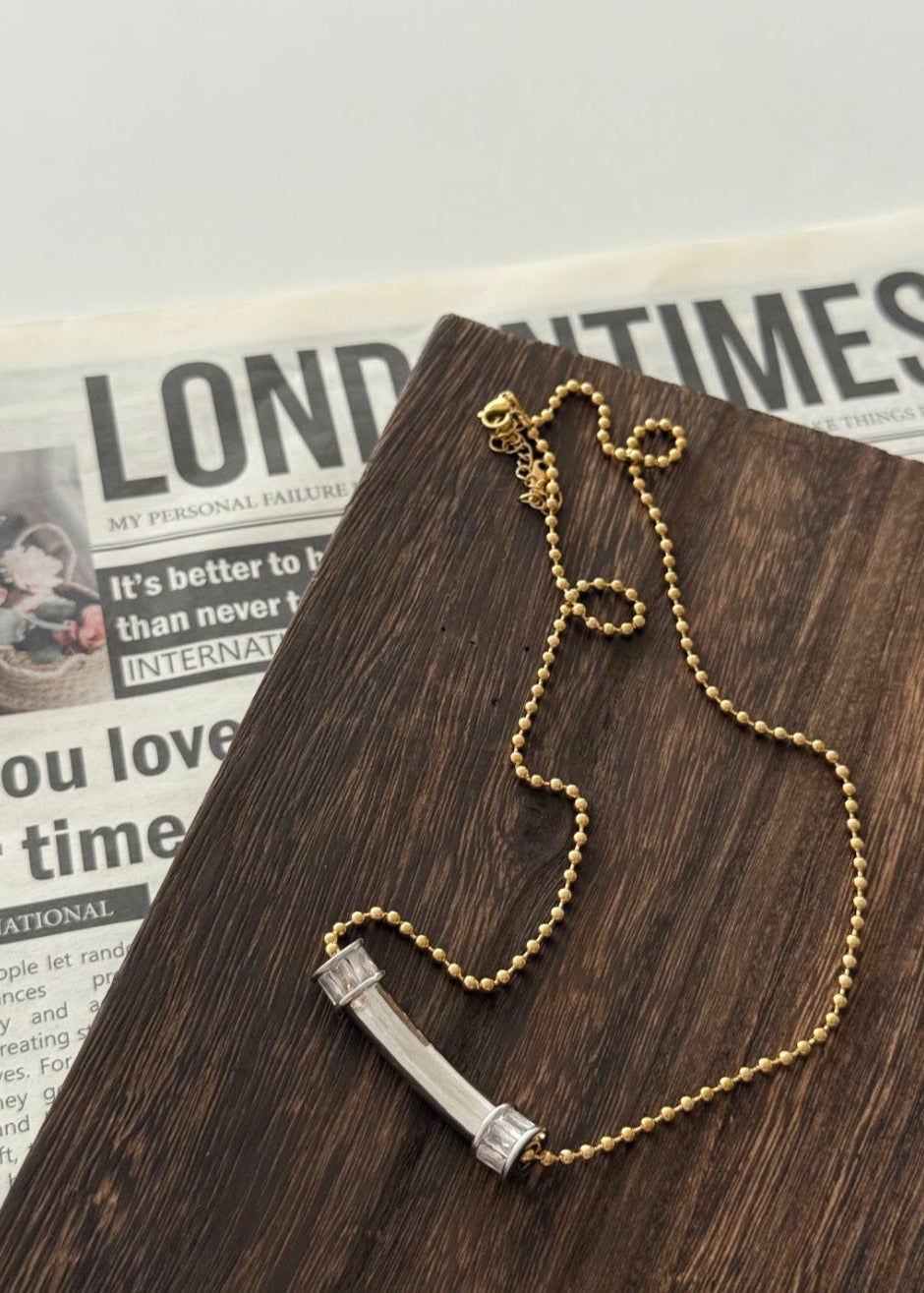 Gold chain necklace with a silver bar pendant on a wooden surface with a newspaper in the background.
