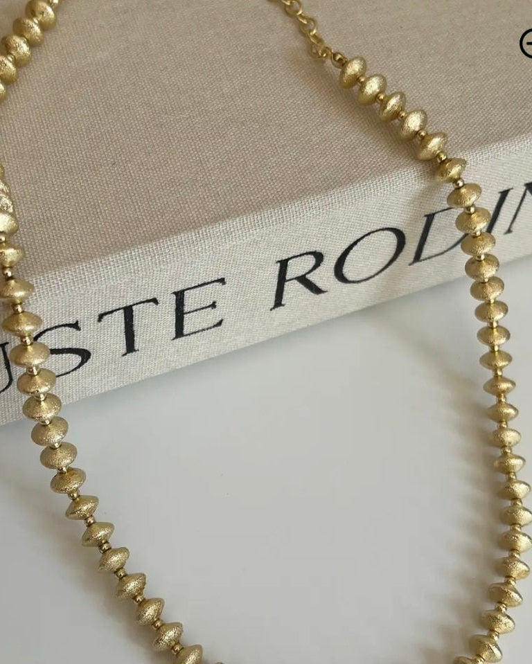 Gold beaded necklace on a beige surface with 'Guste Rodin' branding.