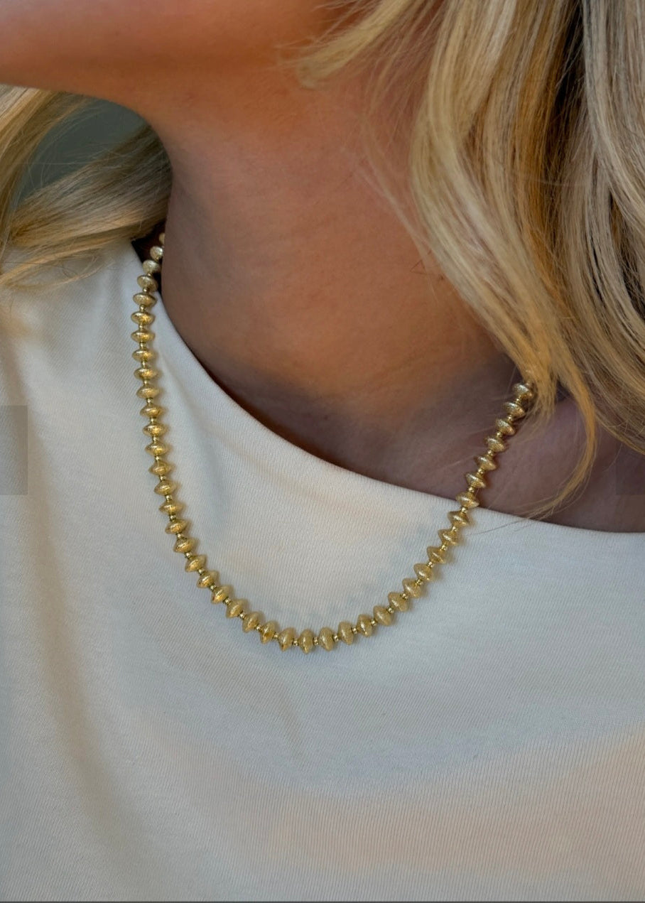 Gold necklace worn by a person with blonde hair, on a plain background